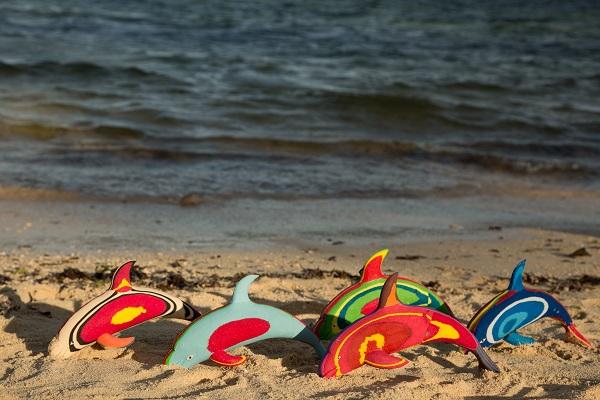 Five hand-carved multicolored dolphin sculptures made of upcycled flip flops by Ocean Sole lined up on the beach by the shoreline.