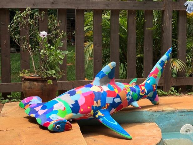 Hand-carved multicolored hammerhead shark sculpture made of upcycled flip flops by Ocean Sole sitting on edge of a pool.