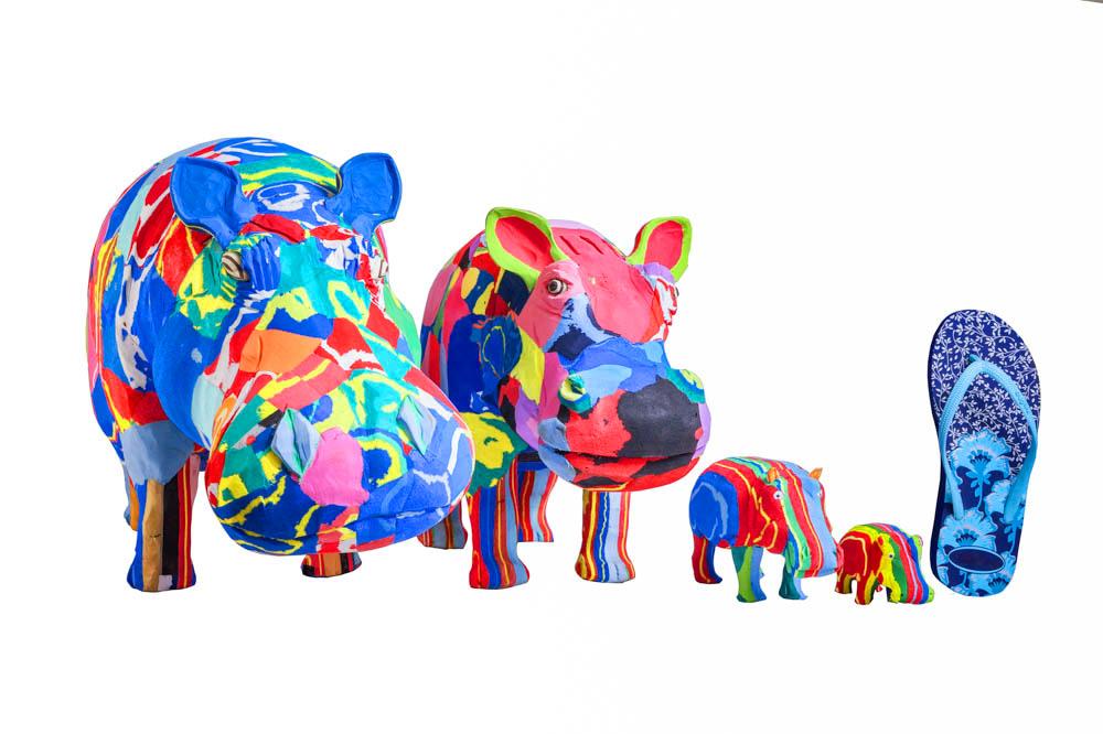 Four hand-carved multicolored hippo sculptures made of upcycled flip flops by Ocean Sole lined up in size comparison to a flip flop.