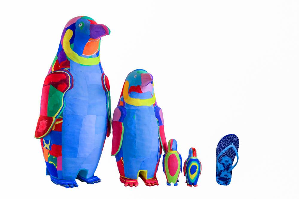Four hand-carved multicolored penguin sculptures made of upcycled flip flops by Ocean Sole lined up in size comparison to a flip flop.