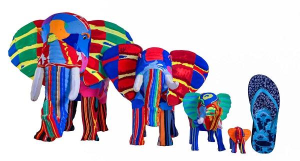 Four hand-carved multicolored elephant sculptures made of upcycled flip flops by Ocean Sole lined up in size comparison to a flip flop.
