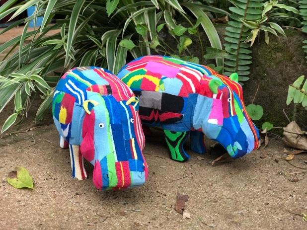 Two hand-carved multicolored hippo sculptures made of upcycled flip flops by Ocean Sole standing outside in greenery.