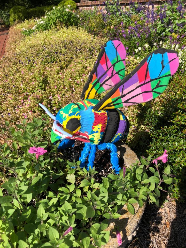 Large multicolored bee sculpture made of upcycled flip flops by Ocean Sole sitting in flower garden.