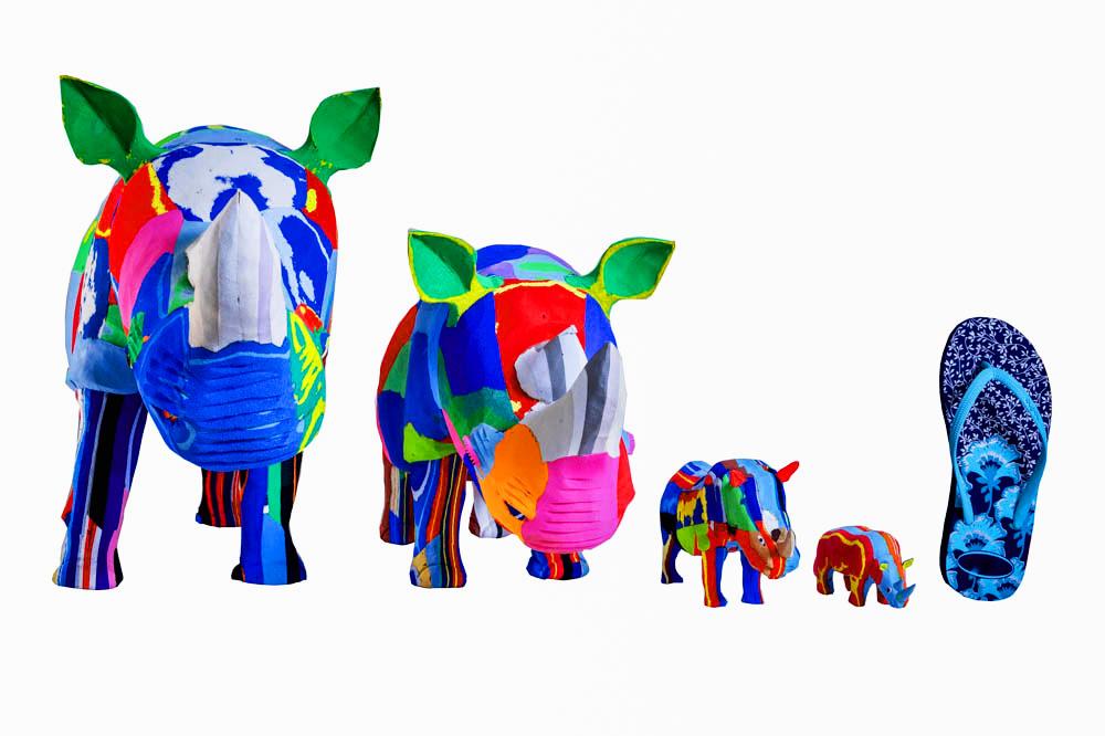 Four hand-carved multicolored rhino sculptures made of upcycled flip flops by Ocean Sole lined up in size comparison to a flip flop.