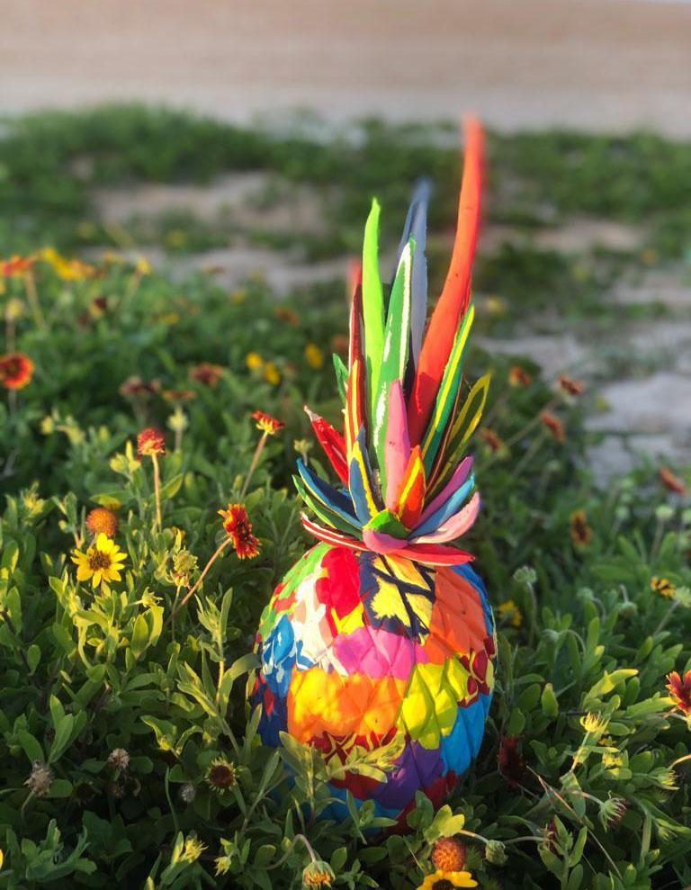 Hand-carved multicolored pineapple sculpture made of upcycled flip flops by Ocean Sole sitting in a flower garden.