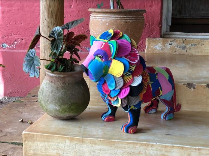 Hand-carved multicolored standing lion sculpture made of upcycled flip flops by Ocean Sole standing on a front porch next to a potted plant. 